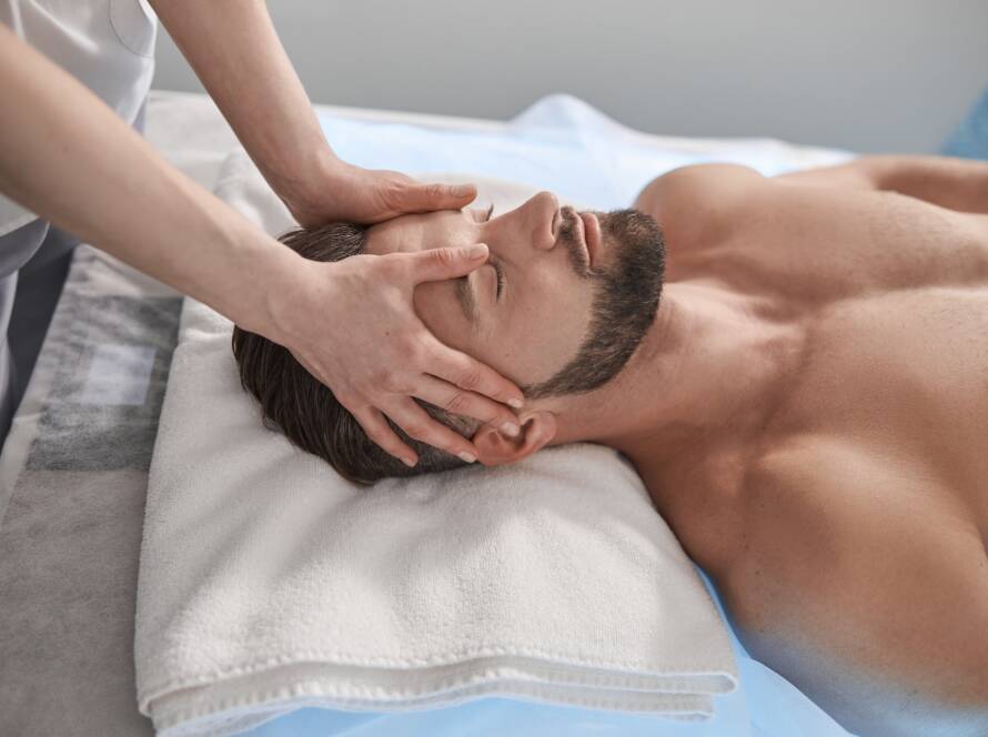 Massage therapist works with head of male lient lying on couch in contemporary clinic